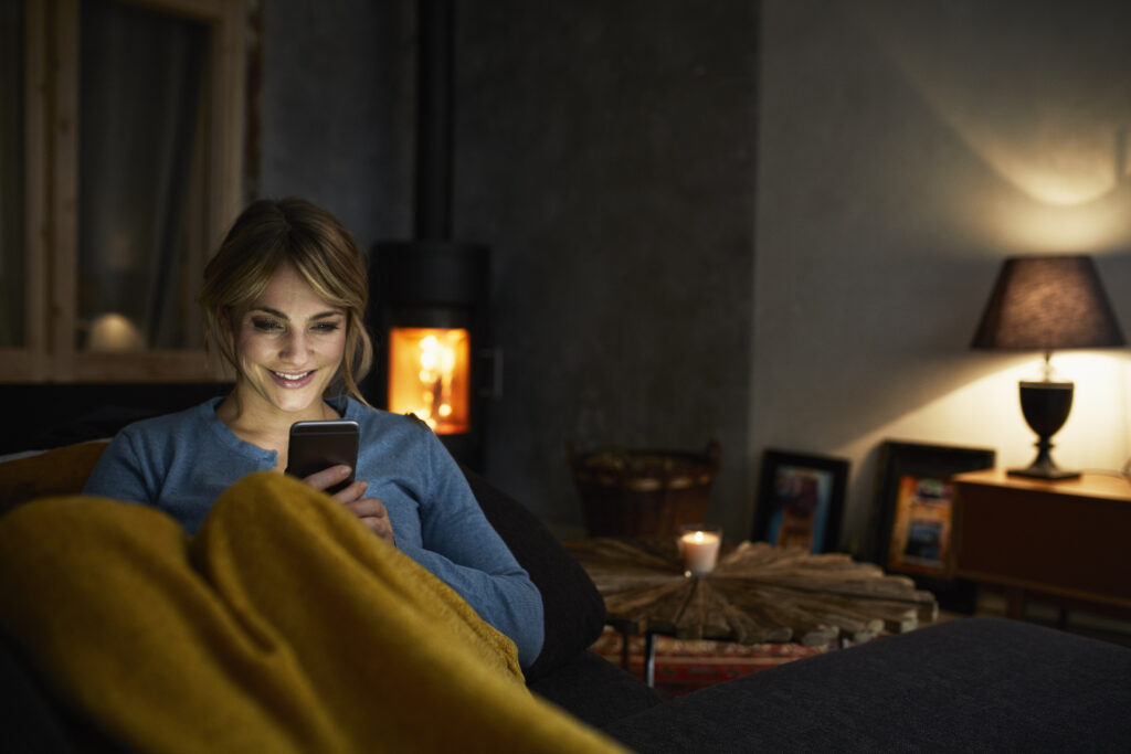 Portrait of smiling woman with smartphone relaxing on couch in the evening - Tinn Energi & Fiber Eva smarthus produkter
